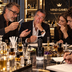A group of people gathered around a polished marble table in a luxury fragrance lounge, smelling and discussing scents. A black and silver VEDLACES 'Royal Decloque' perfume bottle is prominently displayed in the foreground, with other fragrance bottles and glassware around it.