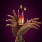 A perfume bottle labeled MON TALE is held between two hands against a dark purple background. The bottle has a gold cap and a gold-toned hanging tag, and the hands are styled with rings and a bracelet.