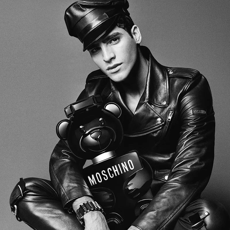 A black and white editorial photo of a model wearing a black leather jacket, matching cap, and trousers, holding a black Moschino teddy bear bottle. The Moschino brand name is clearly visible on the bottle.