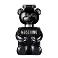 A bottle of Toy Boy Eau de Parfum by Moschino, featuring a black teddy bear shape with a smiling face and holding a sign with the brand name.