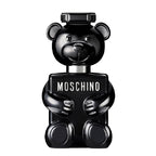 A bottle of Toy Boy Eau de Parfum by Moschino, featuring a black teddy bear shape with a smiling face and holding a sign with the brand name.