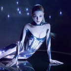 A model wearing a shiny metallic silver bodysuit with integrated arm and leg pieces, posing on a reflective dark surface against a dark background with blue light flares.