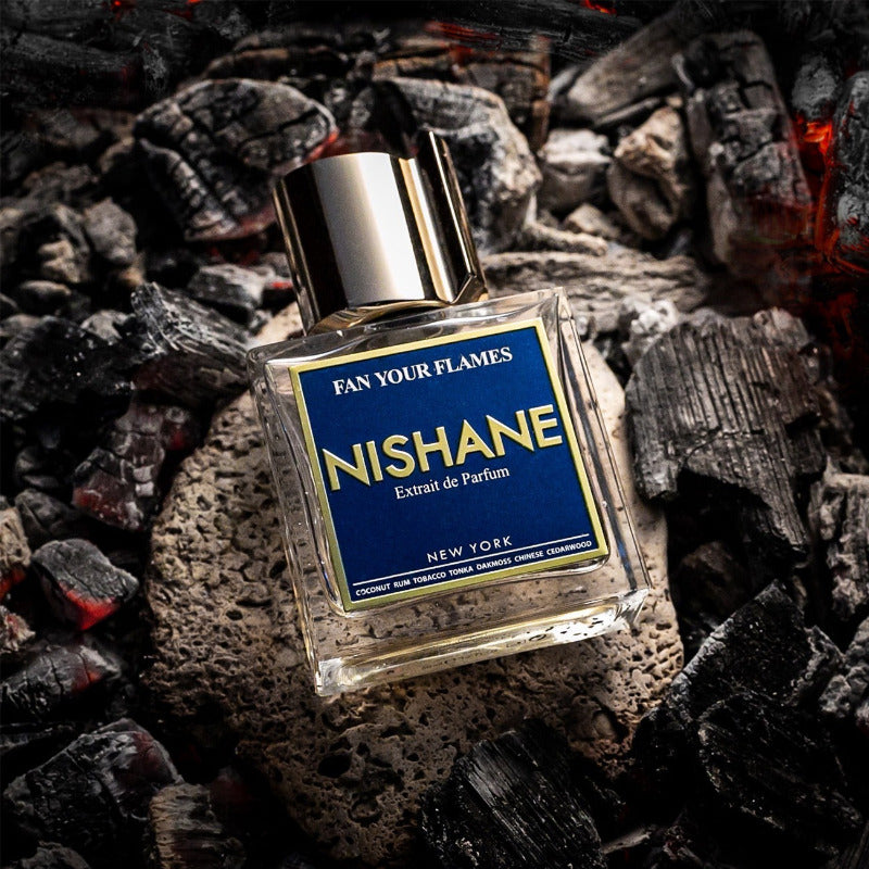 A bottle of Nishane 'Fan Your Flames' Extrait de Parfum placed on a bed of glowing embers and charcoal. The bottle is clear glass with a metallic silver cap and a prominent blue label with gold lettering.