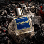 A bottle of Nishane 'Fan Your Flames' Extrait de Parfum placed on a bed of glowing embers and charcoal. The bottle is clear glass with a metallic silver cap and a prominent blue label with gold lettering.