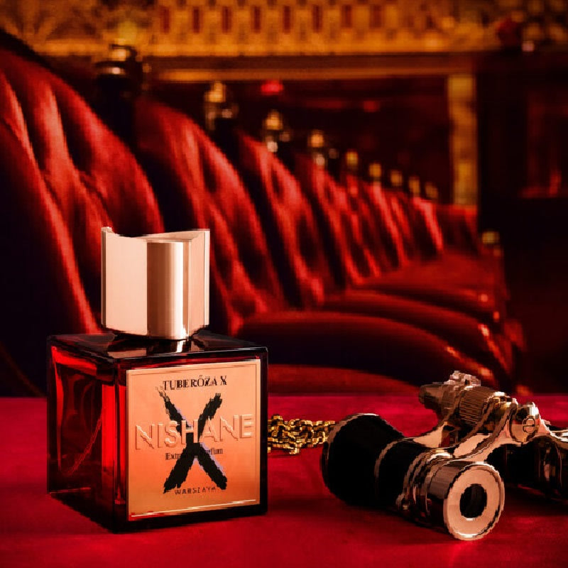 A bottle of Juberogan Nishane Eau de Parfum sits on a red velvet surface with a blurred red theater-style seating background. A vintage-style camera and gold chain are placed beside the bottle as styled props.