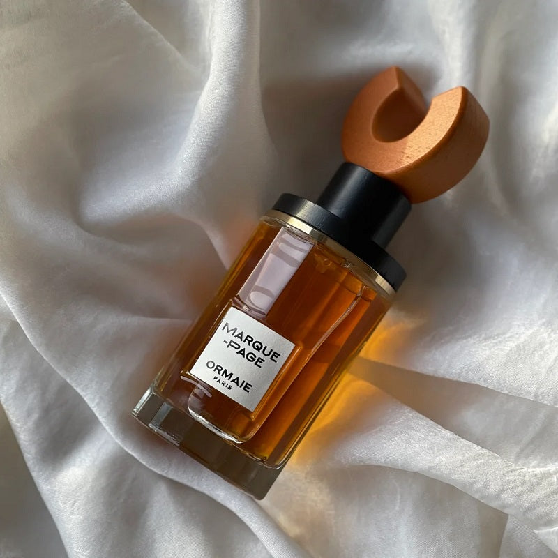 A bottle of Ormaie Marquise d'Or perfume with amber liquid, a black collar, and a distinctive wooden cap, resting on draped white fabric.