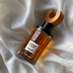 A bottle of Ormaie Marquise d'Or perfume with amber liquid, a black collar, and a distinctive wooden cap, resting on draped white fabric.