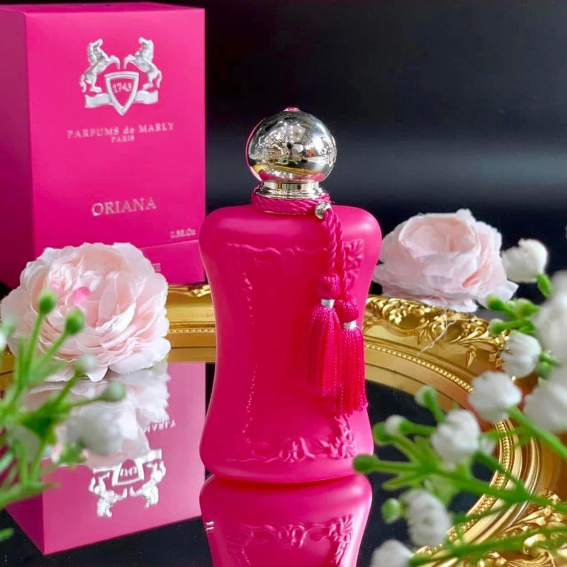 Pink perfume bottle with gold cap on a reflective surface, surrounded by flowers and a pink box labeled 'Oriana'.