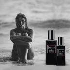 Two black rectangular perfume bottles labeled 'fracas by ROBERT PIGUEZ PARIS' with pink borders on the labels, shown on a beach next to a seated woman in a black-and-white photograph.
