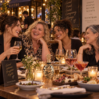 Four women laughing and dining together at a candlelit restaurant table, with Scentbar bottles and signage visible in the scene.