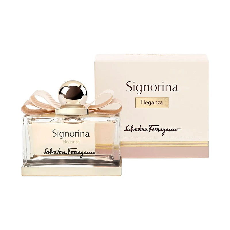 A bottle of Signorina Eleganza Eau de Parfum by Salvatore Ferragamo, with a box in the background. The bottle is clear with a gold cap and a pink ribbon decoration.