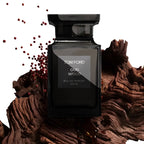 A 100ml bottle of Tom Ford Oud Wood Eau de Parfum, presented in a glossy black glass bottle. The bottle is styled on a textured piece of dark brown wood, with small red berries floating on the left, against a solid white background.