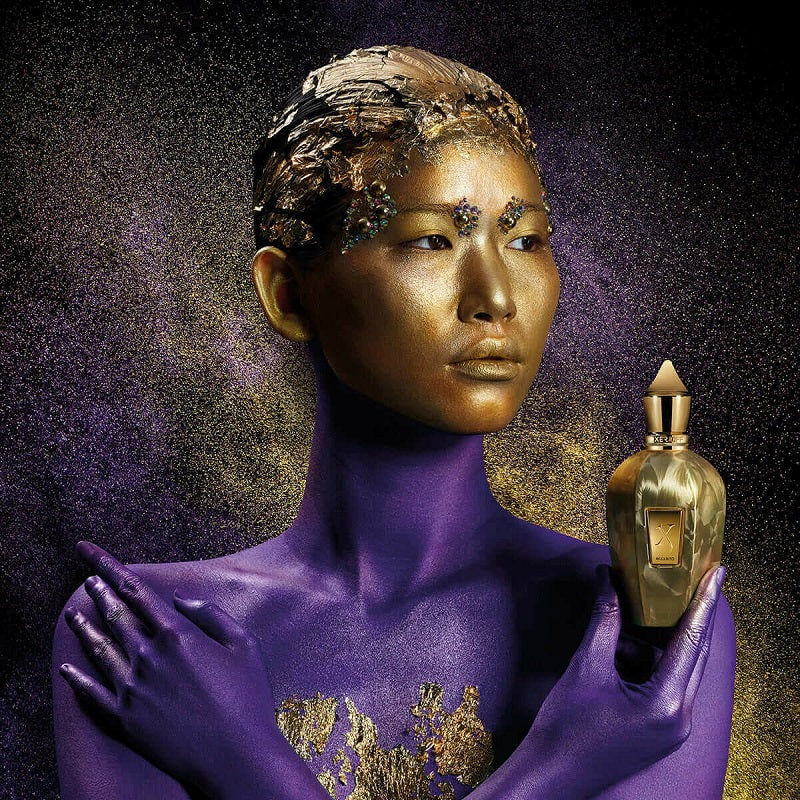 Artistic depiction of a person with gold and purple makeup holding a gold perfume bottle against a dark background.
