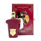 A bottle of Casamorati Italica Eau de Parfum by Xerjoff with a red and gold box in the background. The perfume bottle is clear with a gold cap, and there is a white fabric swatch attached to the box.