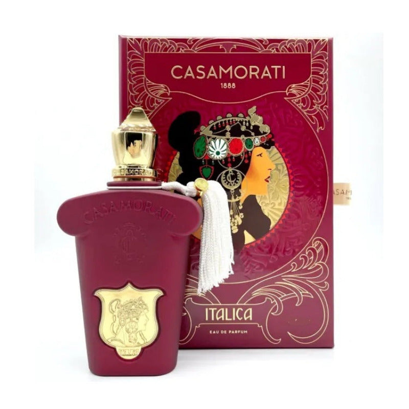 A bottle of Casamorati Italica Eau de Parfum by Xerjoff with a red and gold box in the background. The perfume bottle is clear with a gold cap, and there is a white fabric swatch attached to the box.