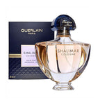 A bottle of Shalimar Cologne Eau de Toilette by Guerlain with a decorative box in the background. The bottle is transparent, revealing a pinkish liquid, with a prominent Guerlain Paris branding.